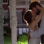A couple embracing and kissing at night in front of a decorative stone structure with columns, with a building and greenery in the background.