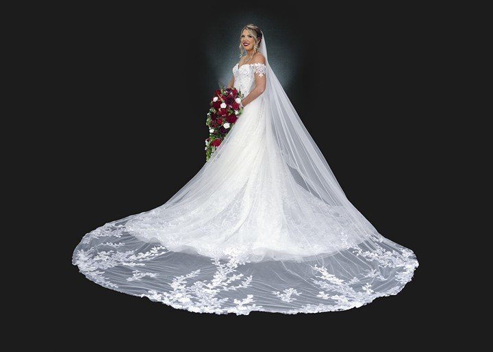 Bridals 1 A bride in a long white wedding gown with a lace train and veil stands smiling for her bridal portrait session, holding a bouquet of red and white flowers against a black background.