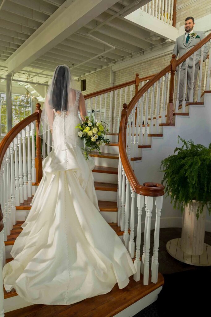Bride in white gown ascending staircase with groom in gray suit at top, indoor setting.