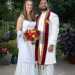 A couple in traditional Indian wedding attire smiles, holding a vibrant bouquet. They're standing outdoors with greenery and flowers in the background, perfectly captured by a talented wedding photographer.
