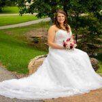 In this beautiful bridal portrait, the bride in a white gown sits gracefully on a stone by the pond, her bouquet of pink and white flowers complementing the lush green grass and trees that frame this idyllic scene.